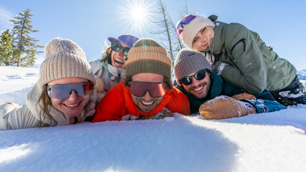 Winter in Flachau (c) Flachau Tourismus/Christian Lorenz Five friends in winter clothes lie smiling in the Flachau snow, with trees and blue sky behind them. (Enlarged view)