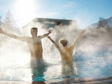 Two people with raised arms enjoy a steaming outdoor pool on a sunny winter’s day, surrounded by mountains.