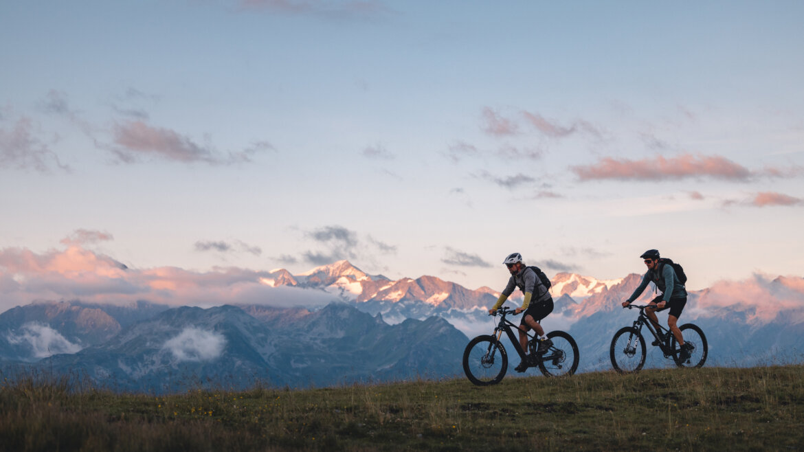 Biken at the Resterkogel(c) Nationalpark Hohe Tauern Mathäus Gartner Two people ride mountain bikes on a grassy hill in National Park Hohe Tauern at sunset with snowy peaks behind. (Enlarged view)