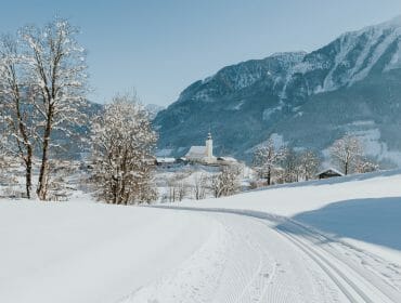 Snowy landscape with trees, a church, and mountains viewed from a sun terrace under clear blue skies.