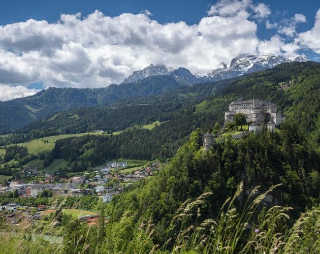A hilltop castle in Werfen overlooks a village and green valley with snow-capped mountains in the background.