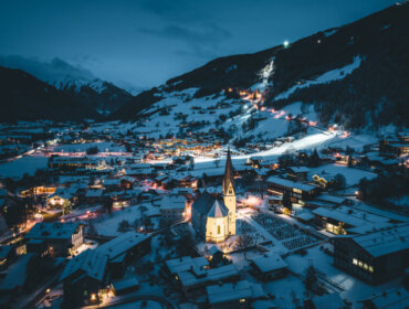 bramberg (c) wildkogelarena Snowy Bramberg mountain village at night, warmly lit buildings, a church, and a glowing ski piste in the background.