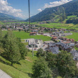 Bramberg im Sommer (c) wildkogelarena Aerial view of Bramberg village, green fields, trees, and mountains under a blue sky with scattered clouds.