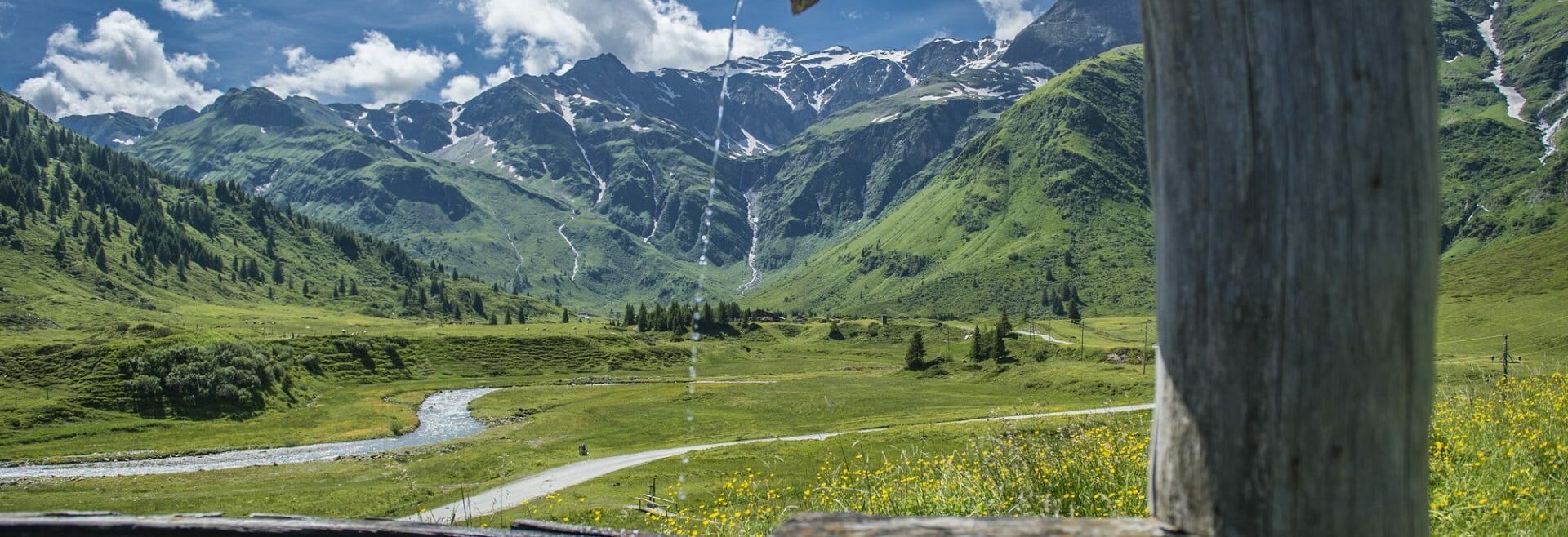 Brunnen im Nassfeld/Sportgastein/Bad Gastein A wooden fountain trickles water in Gastein’s green mountain valley, with a stream and snowy peaks beyond.