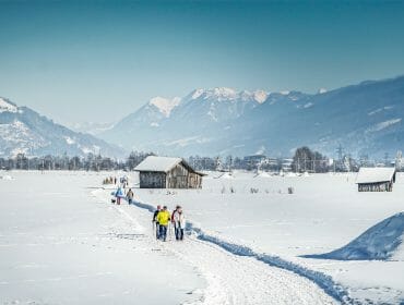 (c) TVB Piesendorf Niedernsill, Harry Liebmann – Winterwandern People cross-country skiing on a snowy trail near Niedernsill, with mountains and wooden cabins in the background.
