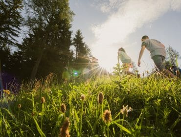 c-tvb-werfenweng_christian-schartner-8_wandern_familie-scaled Children running up a grassy hill in Werfenweng, surrounded by trees and wildflowers under the sun.