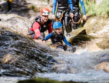An adult and child in helmets and wetsuits slide down a rocky stream in Saalachtal during outdoor adventure.