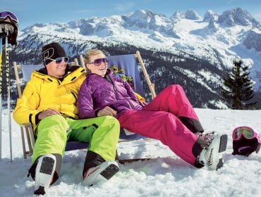 Pärchen in Skikleidung beim Entspannen im Liegestuhl vor winterlicher Kulisse