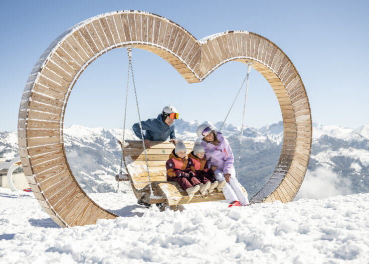 A family sits on a wooden heart-shaped swing in a snowy mountain landscape under a clear sky.