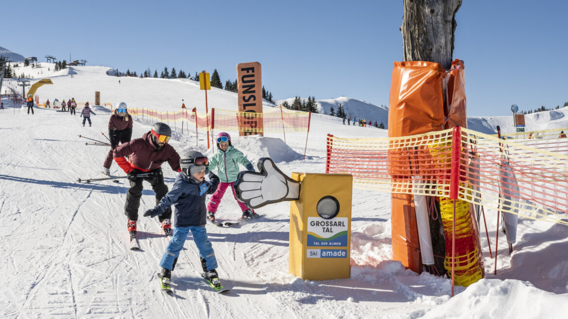 (c) Lorenz Masser Children and adults skiing past a Funslope sign and a thumbs-up marker on a snowy ski slope. (Enlarged view)