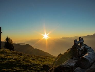 Sunrise over misty mountains with a wooden cross and bench on a grassy hillside.