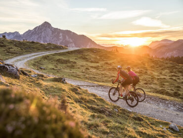 Two cyclists ride a winding mountain trail at sunset in scenic Tennengau region.