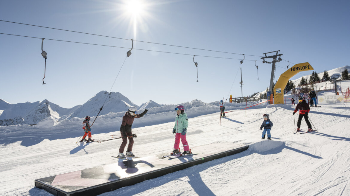 Skiers, including children, using a funslope course on a sunny winter day with snowy mountains in the background. (Enlarged view)