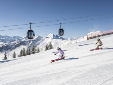 Two people skiing downhill on a snowy slope with ski lifts and mountains in the background.