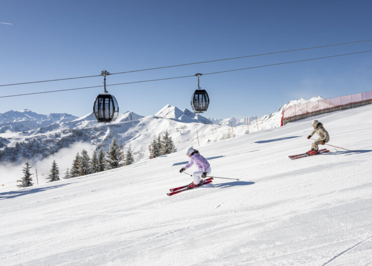 Two people skiing downhill on a snowy slope with ski lifts and mountains in the background.