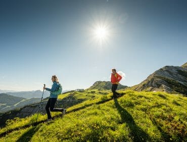 Two people hiking up a grassy mountain in Salzburger Sportwelt under a bright sun and clear blue sky.