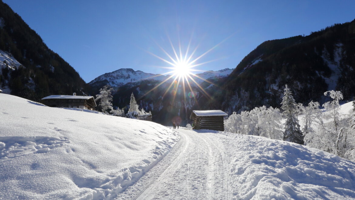 (c) www.grossarltal.info Snow-covered landscape with wooden cabins, trees, mountains, and bright sun shining in a clear blue sky. (Enlarged view)