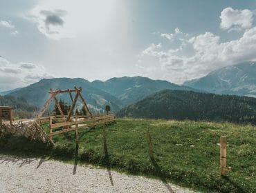 img_9516 Wooden swing set on a grassy hill overlooking forested mountains under a partly cloudy sky.
