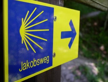 Blau-gelber Wegweiser des Jakobsweges mit der stilisierten Jakobsmuschel als Logo