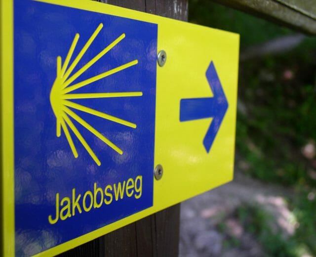 Blau-gelber Wegweiser des Jakobsweges mit der stilisierten Jakobsmuschel als Logo