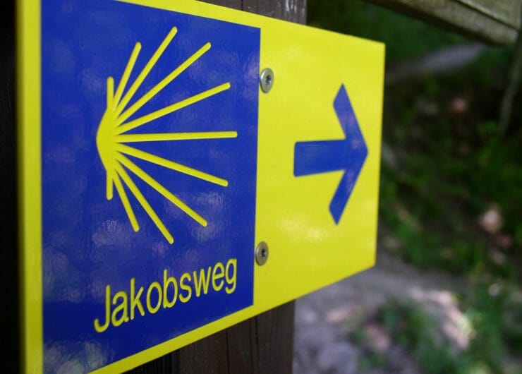 Blau-gelber Wegweiser des Jakobsweges mit der stilisierten Jakobsmuschel als Logo