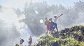 ©Ferienregion Nationalpark Michael Huber A family enjoys hiking near a misty waterfall in National Park Hohe Tauern, surrounded by lush greenery and sunlight.