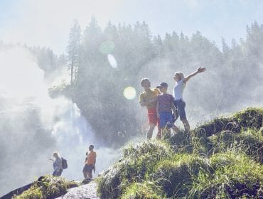 A family enjoys hiking near a misty waterfall in National Park Hohe Tauern, surrounded by lush greenery and sunlight.
