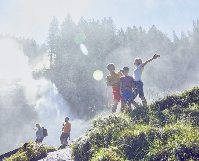 A family enjoys hiking near a misty waterfall in National Park Hohe Tauern, surrounded by lush greenery and sunlight.