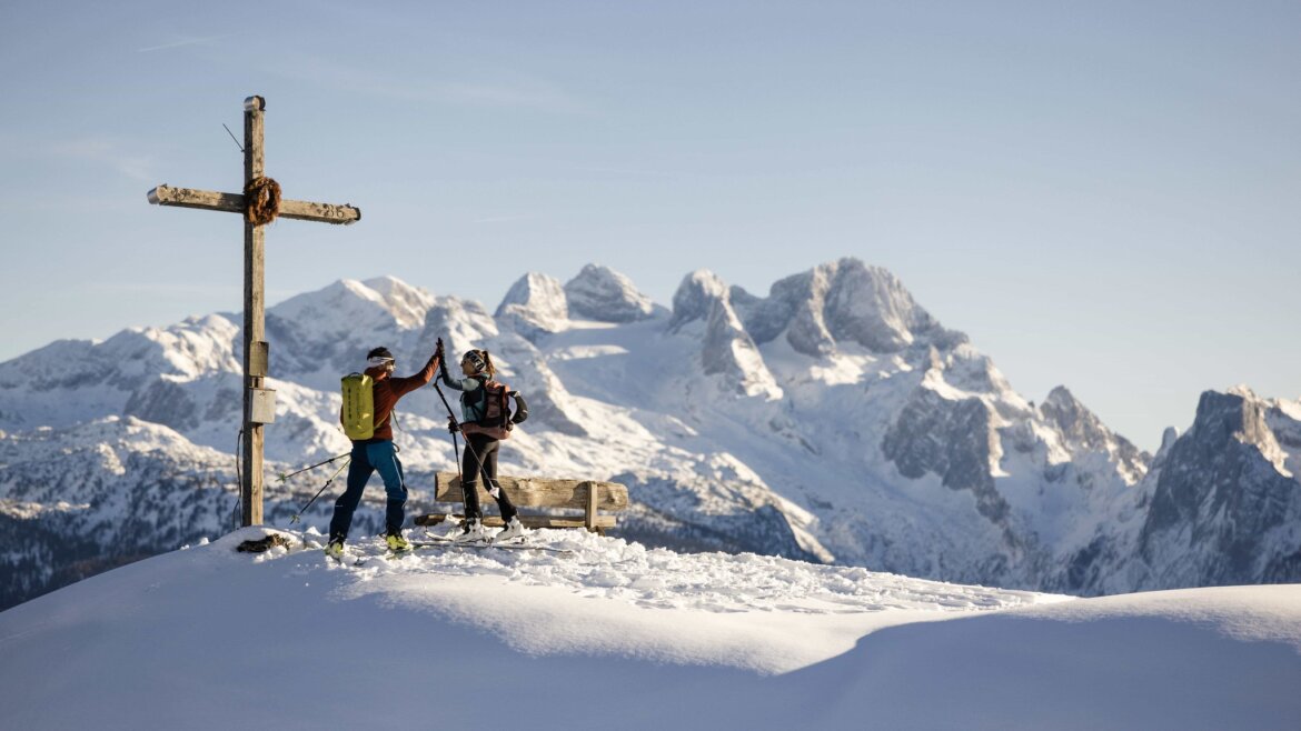 Two people high five beside a wooden cross on a snowy mountain peak in Tennengau.