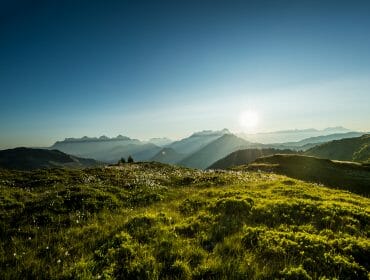 Sunrise over distant mountains in Saalbach Hinterglemm, casting long shadows on a green, grassy hill.