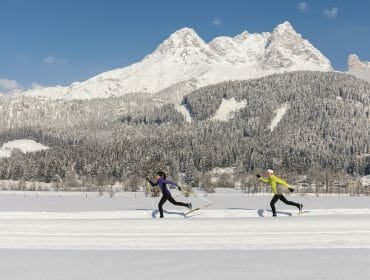 Two people cross-country skiing on a snowy Saalfelden Leogang trail with mountains and pine trees behind.