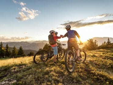 Two cyclists in helmets fist bump at sunset on a grassy Flachau hilltop with mountains in the background.