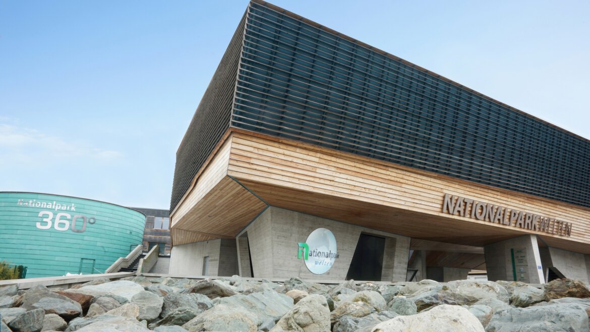 (c) Hohe Tauern Modern building with wooden and metal accents, labelled Nationalpark Hohe Tauern, against rocks and blue sky. (Enlarged view)