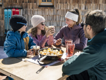 A family in Obertauern enjoys food and drinks outdoors, smiling together after a winter sport adventure.