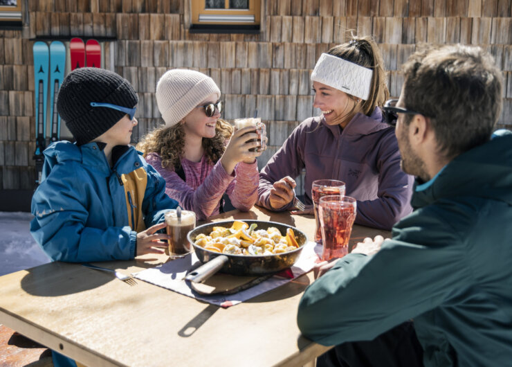 A family in Obertauern enjoys food and drinks outdoors, smiling together after a winter sport adventure.