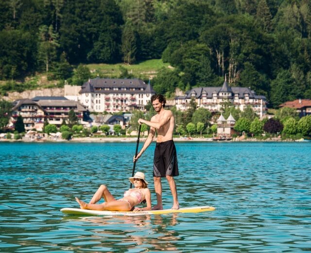 Paerchen am SUP2(c)Salzkammergut A man paddles a woman on a stand-up paddleboard on Fuschlsee, with hotels and trees in the background.