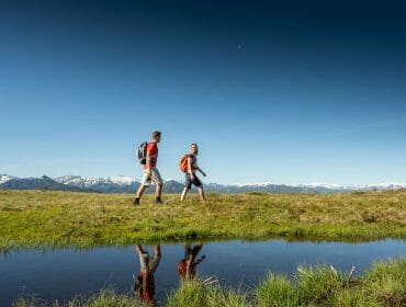Two people walking near Radstadt, mountains and blue sky reflected in a grassy pond.
