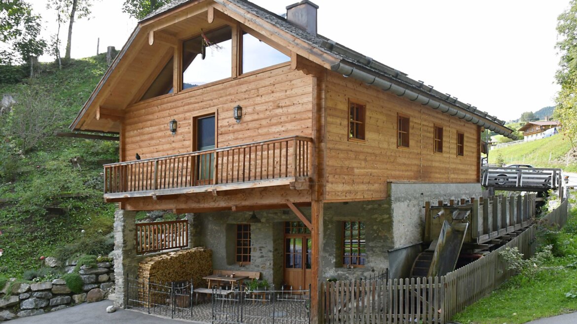 The Rohrmoosschmiede, a two-storey wooden chalet with a balcony, stone foundation, and a stack of firewood outside in a rural setting. (Enlarged view)