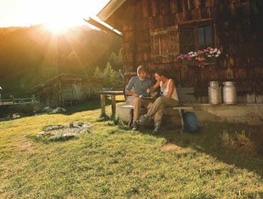 Two people sit outside a rustic cabin at sunset, enjoying mountain scenery—a perfect hut holidays moment.