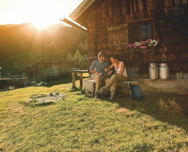 Two people sit outside a rustic cabin at sunset, enjoying mountain scenery—a perfect hut holidays moment.