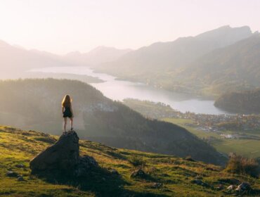 © SalzburgerLand Tourismus, Sebastian Scheichl, Rettenkogel Woman stands on a rock after walking, overlooking a scenic valley with a lake, town, and mountains at sunset.