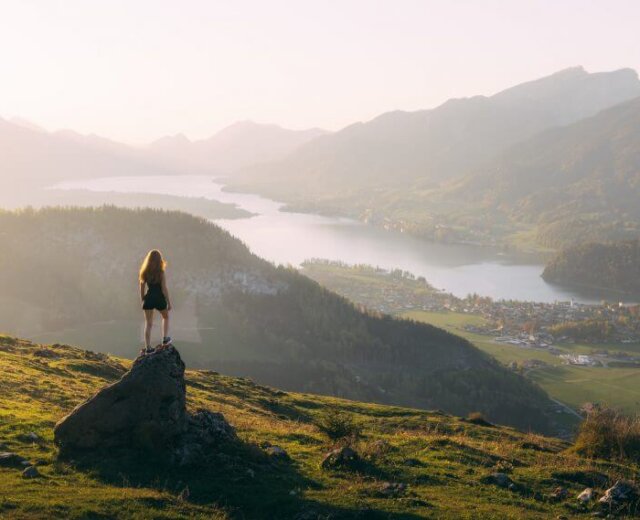 © SalzburgerLand Tourismus, Sebastian Scheichl, Rettenkogel Woman stands on a rock after walking, overlooking a scenic valley with a lake, town, and mountains at sunset.