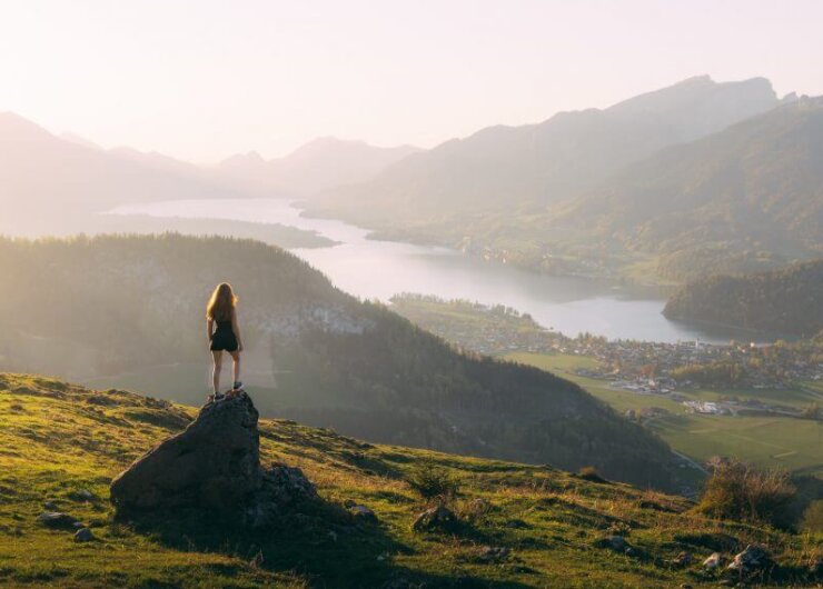 Woman stands on a rock after walking, overlooking a scenic valley with a lake, town, and mountains at sunset.