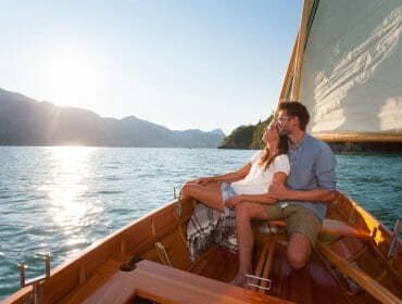 A couple sit in a wooden sailboat, embracing and smiling on sunny Lake St.Gilgen with mountains in the background.