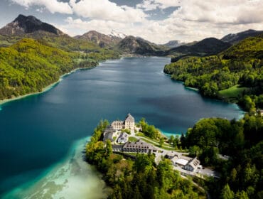 Aerial view of Fuschlsee, a large lake surrounded by green mountains with buildings on its wooded shore.