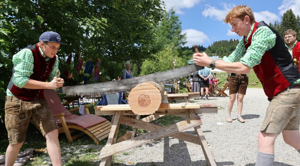 Sawing wood (c) SLT Marcel Müller Two young men in traditional clothes use a two-man saw to cut a log outdoors on a sunny day. (Enlarged view)
