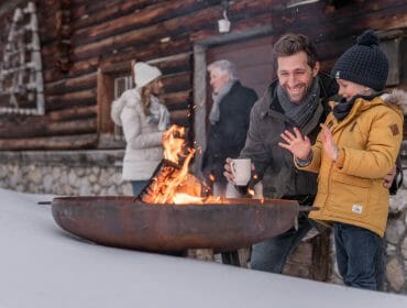 family winter hut A man and child warm up by a fire pit outside a cabin, enjoying cosy hut holidays as others chat nearby.
