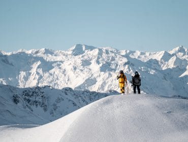 Two people in winter gear stand on a snowy hill in Saalbach Hinterglemm with mountains in the background.