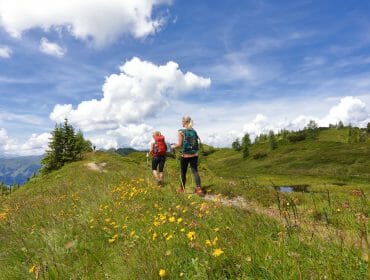 Two people hike on a grassy trail through wildflowers under a blue sky with clouds in the mountains.