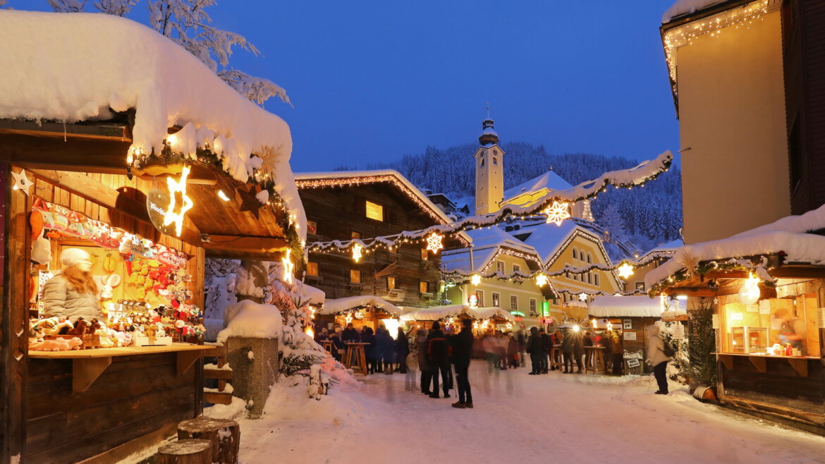 Snowy Christmas market at dusk with wooden stalls, festive lights, and people walking along a decorated street. (Enlarged view)